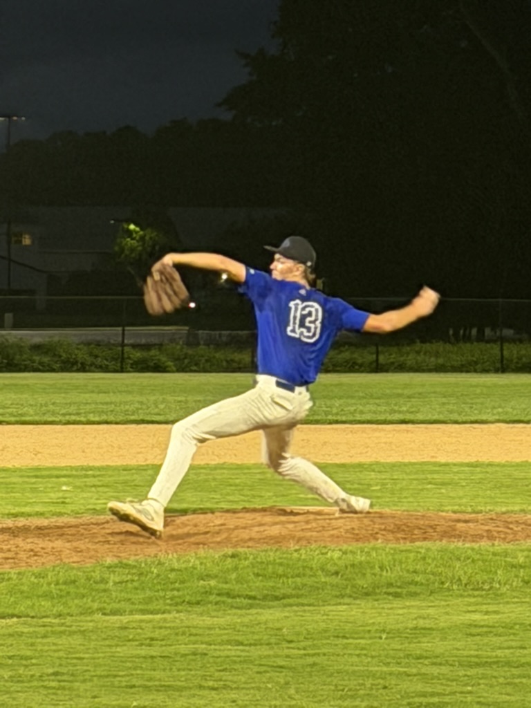 Brayden striding toward home plate during a pitch under the field lights